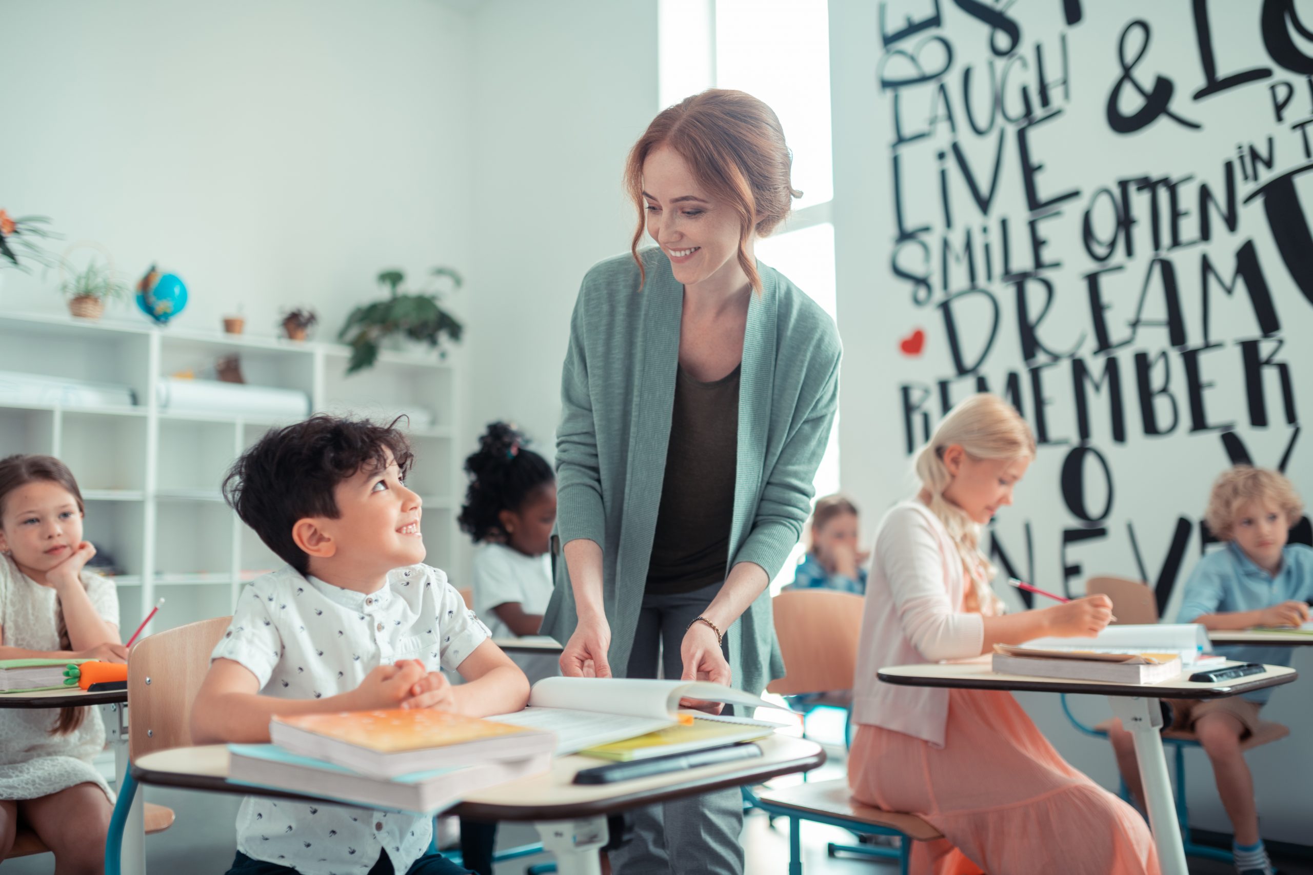 Interacting with pupils. Cheerful pupil sitting at his desk and smiling to his kind teacher standing next to him.